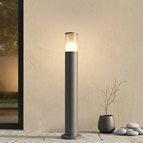 Moderne anthrazitfarbene Pollerleuchte mit warmem Lichtkegel auf einer Steinterrasse vor einer sandbeigen Fassade im minimalistischen Außenbereich.