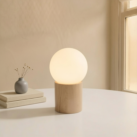 Modern glühende Kugeltischlampe auf hellem Holzsockel in minimalistischem Japandi-Interieur neben einer kleinen Vase und Büchern.